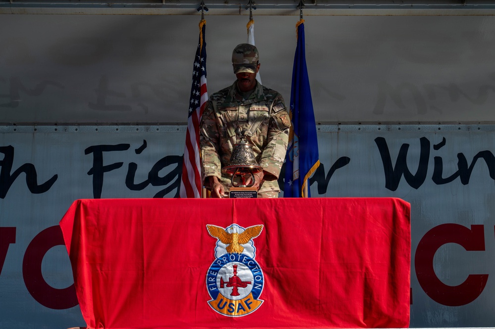 A U.S. Air Force service member in camouflage uniform stands at a podium on a stage, handling a ceremonial bell placed on a red-draped table. The table is covered with a red cloth bearing the USAF Fire Protection insignia. The setting appears to be a formal military ceremony, with the American flag and Air Force flag visible in the background.
