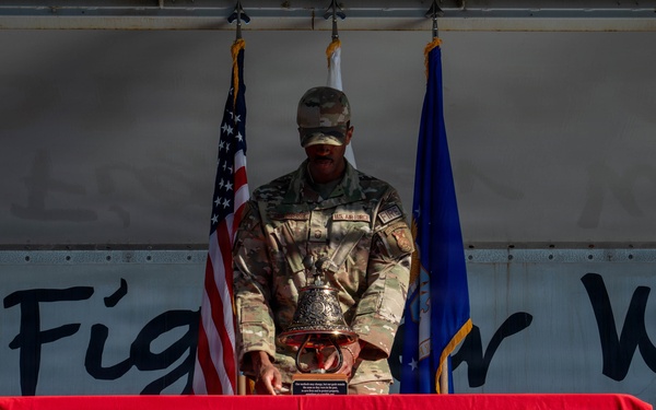 A U.S. Air Force service member in camouflage uniform stands at a podium on a stage, handling a ceremonial bell placed on a red-draped table. The table is covered with a red cloth bearing the USAF Fire Protection insignia. The setting appears to be a formal military ceremony, with the American flag and Air Force flag visible in the background.
