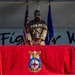 A U.S. Air Force service member in camouflage uniform stands at a podium on a stage, handling a ceremonial bell placed on a red-draped table. The table is covered with a red cloth bearing the USAF Fire Protection insignia. The setting appears to be a formal military ceremony, with the American flag and Air Force flag visible in the background.