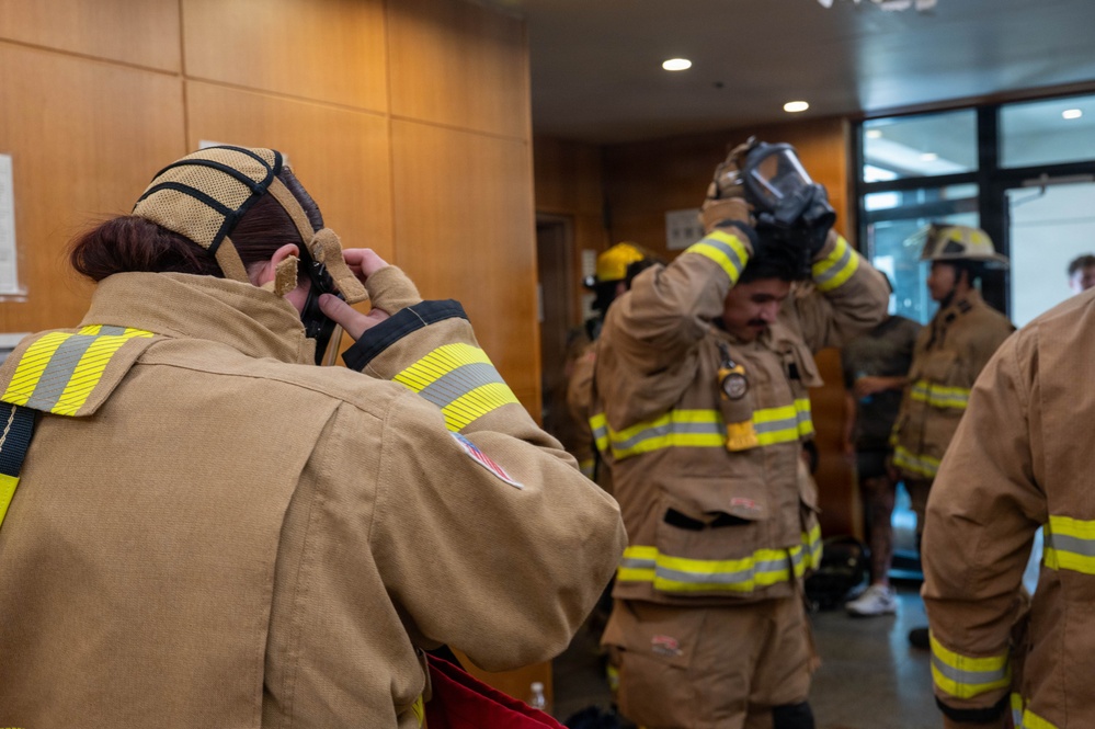 A group of firefighters in tan/khaki turnout gear with yellow-green reflective stripes are donning their equipment inside what appears to be a building lobby or training facility. The foreground shows a female firefighter putting on her hood, while a male firefighter in the middle is lifting a respirator/SCBA mask over his head. The setting suggests a pre-deployment preparation or training exercise.