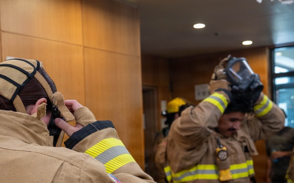 A group of firefighters in tan/khaki turnout gear with yellow-green reflective stripes are donning their equipment inside what appears to be a building lobby or training facility. The foreground shows a female firefighter putting on her hood, while a male firefighter in the middle is lifting a respirator/SCBA mask over his head. The setting suggests a pre-deployment preparation or training exercise.