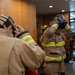 A group of firefighters in tan/khaki turnout gear with yellow-green reflective stripes are donning their equipment inside what appears to be a building lobby or training facility. The foreground shows a female firefighter putting on her hood, while a male firefighter in the middle is lifting a respirator/SCBA mask over his head. The setting suggests a pre-deployment preparation or training exercise.