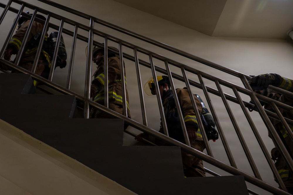 Multiple firefighters in full turnout gear are ascending an interior stairwell, viewed from a low angle looking upward. They are equipped with SCBA (Self-Contained Breathing Apparatus) tanks and wearing standard firefighting protective clothing with yellow reflective striping. The scene appears to depict a training exercise or active emergency response inside a building.