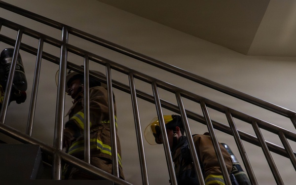 Multiple firefighters in full turnout gear are ascending an interior stairwell, viewed from a low angle looking upward. They are equipped with SCBA (Self-Contained Breathing Apparatus) tanks and wearing standard firefighting protective clothing with yellow reflective striping. The scene appears to depict a training exercise or active emergency response inside a building.
