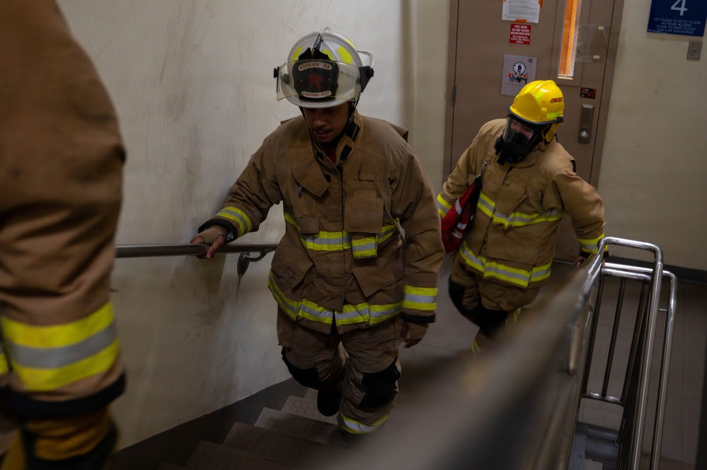 Two firefighters in full turnout gear are ascending a stairwell inside a building, likely during a training exercise or emergency response. The lead firefighter wears a white helmet and tan bunker gear with reflective yellow stripes, while the second wears a yellow helmet and breathing apparatus. A floor number '4' sign is visible on the door in the background.
