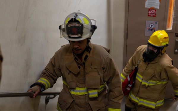 Two firefighters in full turnout gear are ascending a stairwell inside a building, likely during a training exercise or emergency response. The lead firefighter wears a white helmet and tan bunker gear with reflective yellow stripes, while the second wears a yellow helmet and breathing apparatus. A floor number '4' sign is visible on the door in the background.