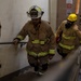 Two firefighters in full turnout gear are ascending a stairwell inside a building, likely during a training exercise or emergency response. The lead firefighter wears a white helmet and tan bunker gear with reflective yellow stripes, while the second wears a yellow helmet and breathing apparatus. A floor number '4' sign is visible on the door in the background.