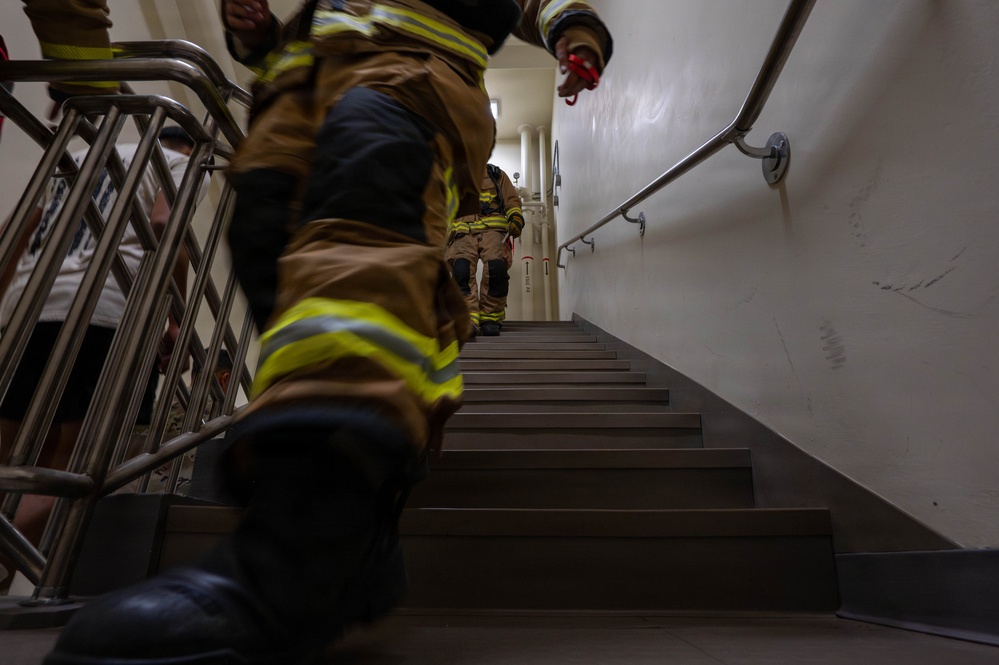 The image shows firefighters in full turnout gear climbing a stairwell, photographed from a low angle looking upward. At least two firefighters are visible wearing tan/brown bunker gear with yellow reflective stripes. This appears to be a stair climb event or training exercise, possibly a memorial stair climb.