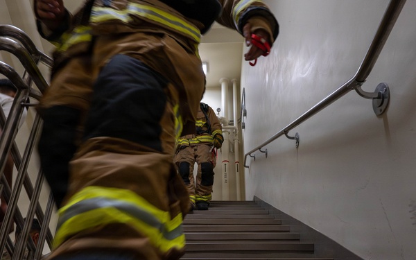 The image shows firefighters in full turnout gear climbing a stairwell, photographed from a low angle looking upward. At least two firefighters are visible wearing tan/brown bunker gear with yellow reflective stripes. This appears to be a stair climb event or training exercise, possibly a memorial stair climb.