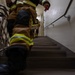 The image shows firefighters in full turnout gear climbing a stairwell, photographed from a low angle looking upward. At least two firefighters are visible wearing tan/brown bunker gear with yellow reflective stripes. This appears to be a stair climb event or training exercise, possibly a memorial stair climb.