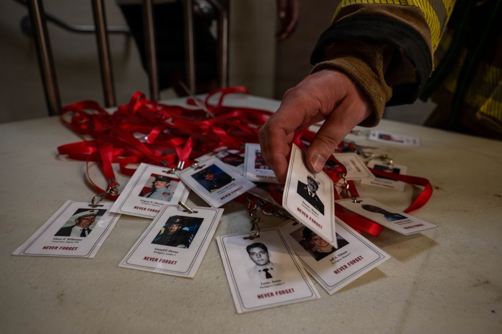 A person wearing a firefighter's jacket is handling memorial lanyards spread across a white table. The lanyards feature photographs and names of individuals with the text 'NEVER FORGET,' suggesting these are commemorative badges honoring fallen firefighters. The red lanyards and formal portrait-style photos indicate this is a memorial or tribute event.