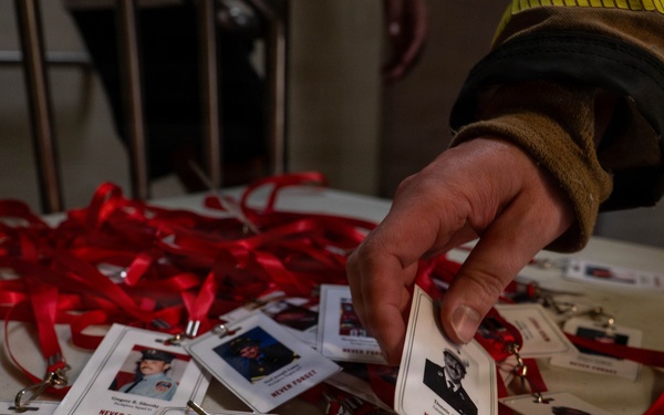 A person wearing a firefighter's jacket is handling memorial lanyards spread across a white table. The lanyards feature photographs and names of individuals with the text 'NEVER FORGET,' suggesting these are commemorative badges honoring fallen firefighters. The red lanyards and formal portrait-style photos indicate this is a memorial or tribute event.
