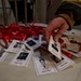 A person wearing a firefighter's jacket is handling memorial lanyards spread across a white table. The lanyards feature photographs and names of individuals with the text 'NEVER FORGET,' suggesting these are commemorative badges honoring fallen firefighters. The red lanyards and formal portrait-style photos indicate this is a memorial or tribute event.