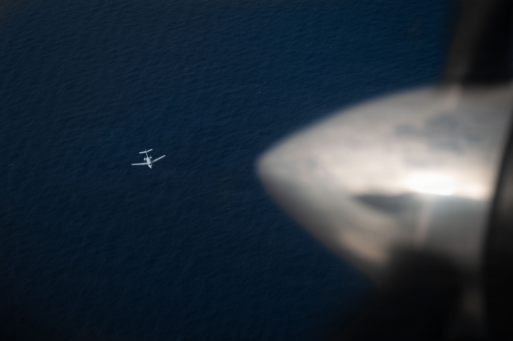 An aerial photograph taken from inside one aircraft looking down at a second, smaller aircraft flying over a large body of dark blue water. The foreground shows a blurred engine nacelle or wingtip of the photographing aircraft. The distant aircraft appears to be a small twin-engine or propeller plane flying at a lower altitude.