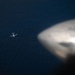 An aerial photograph taken from inside one aircraft looking down at a second, smaller aircraft flying over a large body of dark blue water. The foreground shows a blurred engine nacelle or wingtip of the photographing aircraft. The distant aircraft appears to be a small twin-engine or propeller plane flying at a lower altitude.