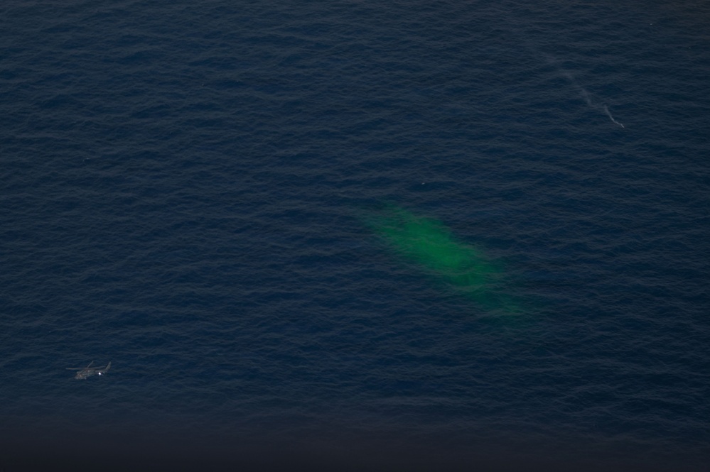 An aerial photograph of open ocean showing a distinctive bright green patch on the water surface, likely a dye marker used in search and rescue or military operations. A small vessel or aircraft is visible in the lower left portion of the image. The image appears to document an active maritime operation or emergency response scenario.
