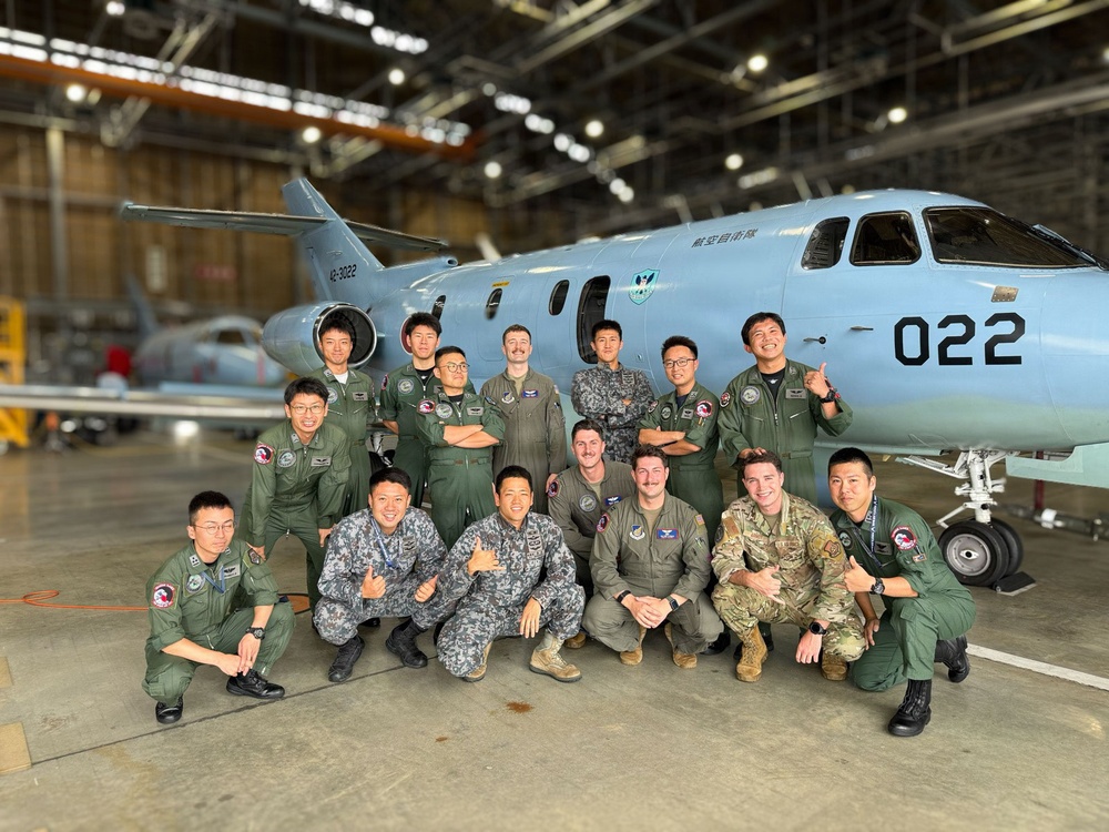 A group of approximately 16 military personnel pose for a group photo inside an aircraft hangar in front of a light blue military aircraft marked '022' and bearing Japanese text '航空自衛隊' (Japan Air Self-Defense Force). The group appears to include both Japanese Air Self-Defense Force members and what appear to be U.S. military personnel in various uniforms, suggesting a joint military exchange or training event. The setting is a well-lit military hangar with industrial overhead structure visible.