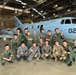 A group of approximately 16 military personnel pose for a group photo inside an aircraft hangar in front of a light blue military aircraft marked '022' and bearing Japanese text '航空自衛隊' (Japan Air Self-Defense Force). The group appears to include both Japanese Air Self-Defense Force members and what appear to be U.S. military personnel in various uniforms, suggesting a joint military exchange or training event. The setting is a well-lit military hangar with industrial overhead structure visible.