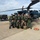 A group of approximately ten military personnel in flight suits pose for a group photo in front of a dark-colored UH-60 Black Hawk helicopter on a tarmac. The group appears to include both Japanese Air Self-Defense Force members and at least two Western (likely U.S.) military personnel, suggesting a joint military exercise or exchange program. Ground crew members are visible on top of and around the helicopter, apparently conducting maintenance or post-flight checks.