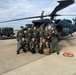 A group of approximately ten military personnel in flight suits pose for a group photo in front of a dark-colored UH-60 Black Hawk helicopter on a tarmac. The group appears to include both Japanese Air Self-Defense Force members and at least two Western (likely U.S.) military personnel, suggesting a joint military exercise or exchange program. Ground crew members are visible on top of and around the helicopter, apparently conducting maintenance or post-flight checks.
