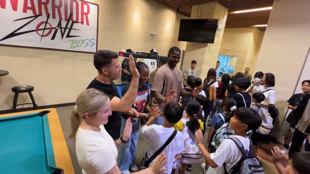 A group of adults and children are gathered inside a facility called 'Warrior Zone BOSS,' engaging in high-fives and friendly interaction. The scene appears to be a community or youth outreach event where adults are welcoming a group of children, many of whom are wearing backpacks. The atmosphere is energetic and celebratory.