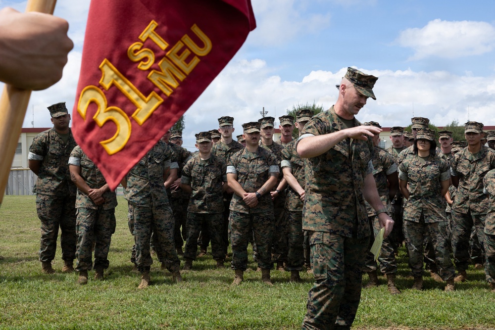 A group of U.S. Marines in woodland MARPAT camouflage uniforms are assembled in formation on a grass field. A senior Marine, likely an officer or senior NCO, appears to be addressing the formation while holding a document. A guidon bearing the '31st MEU' (Marine Expeditionary Unit) designation is prominently displayed in the foreground.