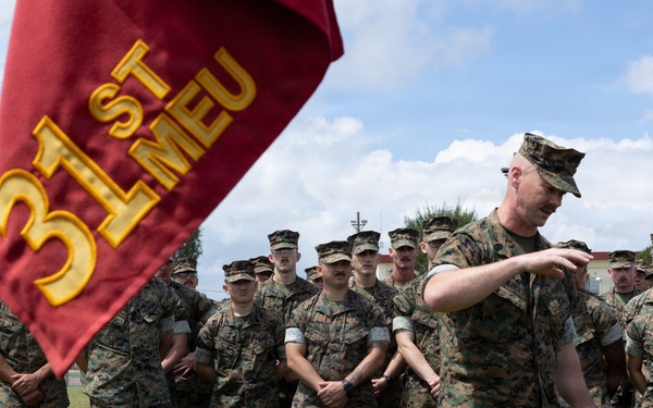 A group of U.S. Marines in woodland MARPAT camouflage uniforms are assembled in formation on a grass field. A senior Marine, likely an officer or senior NCO, appears to be addressing the formation while holding a document. A guidon bearing the '31st MEU' (Marine Expeditionary Unit) designation is prominently displayed in the foreground.