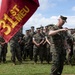 A group of U.S. Marines in woodland MARPAT camouflage uniforms are assembled in formation on a grass field. A senior Marine, likely an officer or senior NCO, appears to be addressing the formation while holding a document. A guidon bearing the '31st MEU' (Marine Expeditionary Unit) designation is prominently displayed in the foreground.