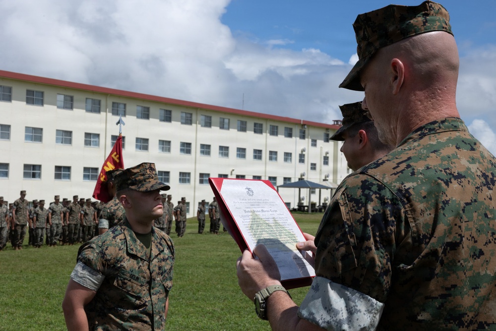 A United States Marine Corps military ceremony is taking place on a parade ground, where a senior Marine is reading from an official document in a red folder to a junior Marine standing at attention. The document is visibly headed 'United States Marine Corps,' suggesting this is likely a promotion, award, or commendation ceremony. A formation of Marines stands in the background alongside a USMC unit guidon flag.