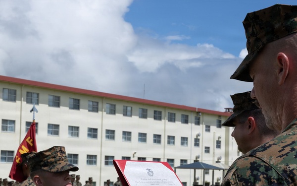 A United States Marine Corps military ceremony is taking place on a parade ground, where a senior Marine is reading from an official document in a red folder to a junior Marine standing at attention. The document is visibly headed 'United States Marine Corps,' suggesting this is likely a promotion, award, or commendation ceremony. A formation of Marines stands in the background alongside a USMC unit guidon flag.