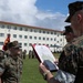 A United States Marine Corps military ceremony is taking place on a parade ground, where a senior Marine is reading from an official document in a red folder to a junior Marine standing at attention. The document is visibly headed 'United States Marine Corps,' suggesting this is likely a promotion, award, or commendation ceremony. A formation of Marines stands in the background alongside a USMC unit guidon flag.