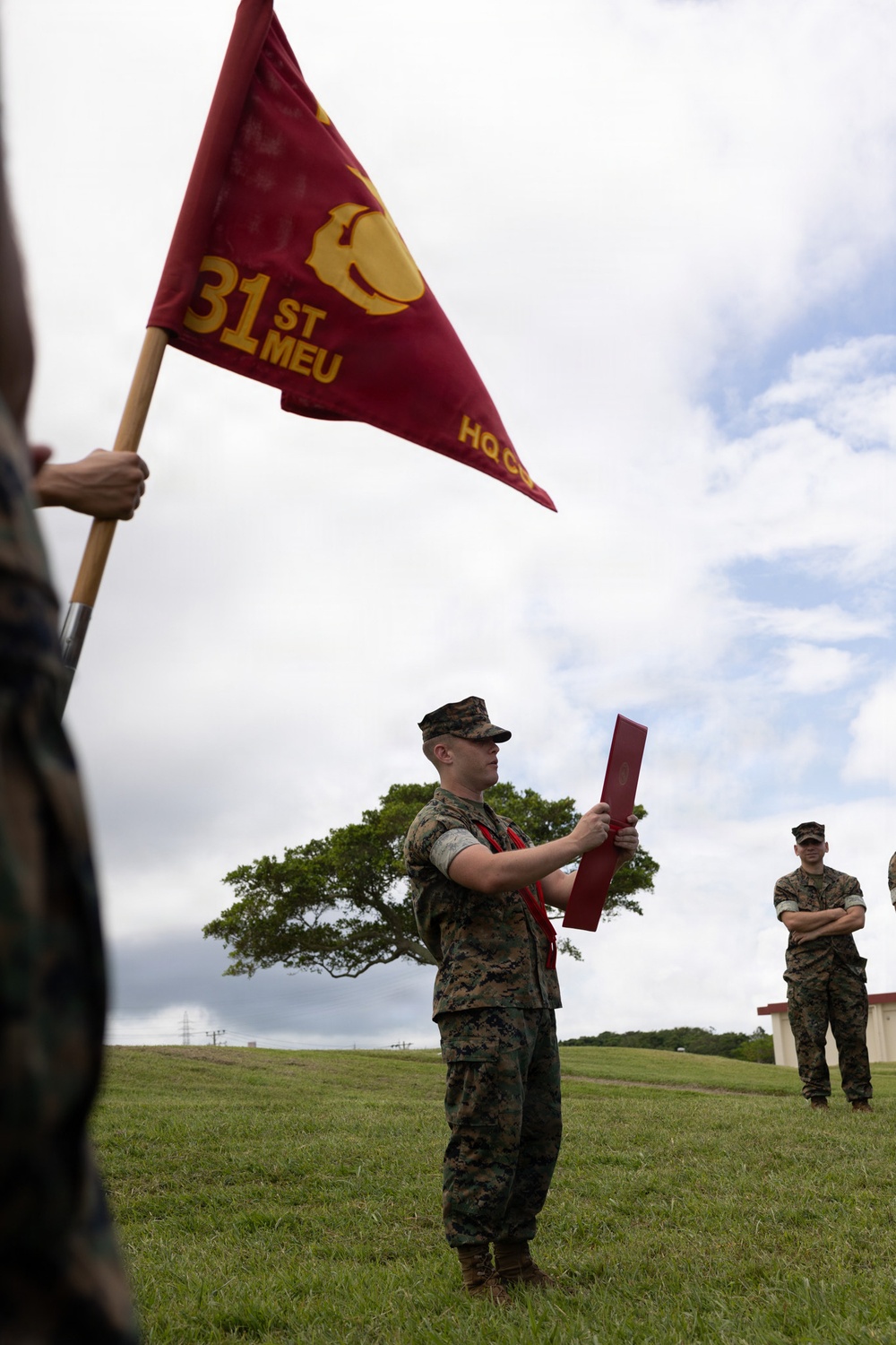 A U.S. Marine in MARPAT camouflage uniform holds a red folder/document during what appears to be a military ceremony or formation outdoors. A red pennant flag bearing '31st MEU' and 'HQ CO' with the Marine Corps Eagle, Globe, and Anchor emblem is prominently displayed in the foreground. The event takes place on a grass field, likely at a military installation in the Pacific region.