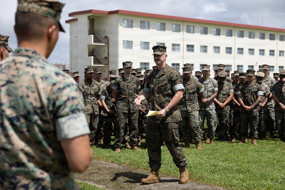 A military formation is taking place outdoors on a grass field in front of a multi-story white building with red trim, consistent with a military barracks or administrative facility. A central figure in U.S. Marine Corps MARPAT camouflage uniform is addressing the assembled troops while holding papers, appearing to be a senior NCO or officer conducting a briefing or formation. A large group of Marines stands in organized rows in the background.