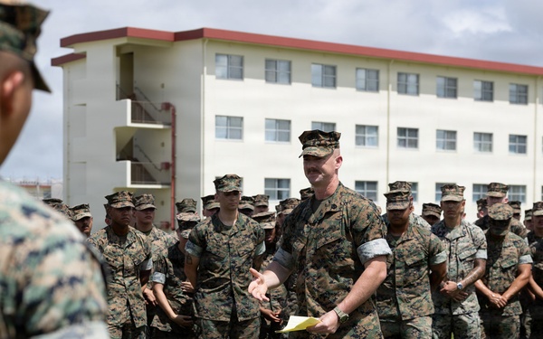 A military formation is taking place outdoors on a grass field in front of a multi-story white building with red trim, consistent with a military barracks or administrative facility. A central figure in U.S. Marine Corps MARPAT camouflage uniform is addressing the assembled troops while holding papers, appearing to be a senior NCO or officer conducting a briefing or formation. A large group of Marines stands in organized rows in the background.