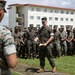 A military formation is taking place outdoors on a grass field in front of a multi-story white building with red trim, consistent with a military barracks or administrative facility. A central figure in U.S. Marine Corps MARPAT camouflage uniform is addressing the assembled troops while holding papers, appearing to be a senior NCO or officer conducting a briefing or formation. A large group of Marines stands in organized rows in the background.