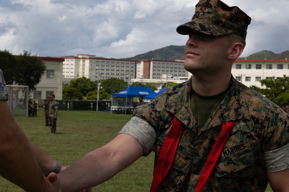 A U.S. Marine in MARPAT digital camouflage uniform is receiving what appears to be a blood pressure check or medical assessment on an outdoor field. The Marine is wearing a red sash/lanyard and has a medical cuff on their arm. The setting appears to be a military installation with large barracks-style buildings visible in the background.