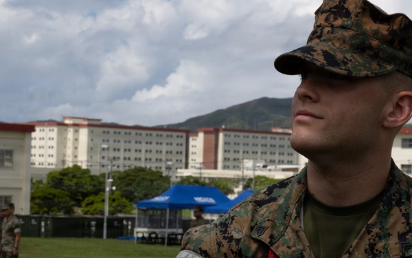 A U.S. Marine in MARPAT digital camouflage uniform is receiving what appears to be a blood pressure check or medical assessment on an outdoor field. The Marine is wearing a red sash/lanyard and has a medical cuff on their arm. The setting appears to be a military installation with large barracks-style buildings visible in the background.