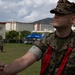 A U.S. Marine in MARPAT digital camouflage uniform is receiving what appears to be a blood pressure check or medical assessment on an outdoor field. The Marine is wearing a red sash/lanyard and has a medical cuff on their arm. The setting appears to be a military installation with large barracks-style buildings visible in the background.