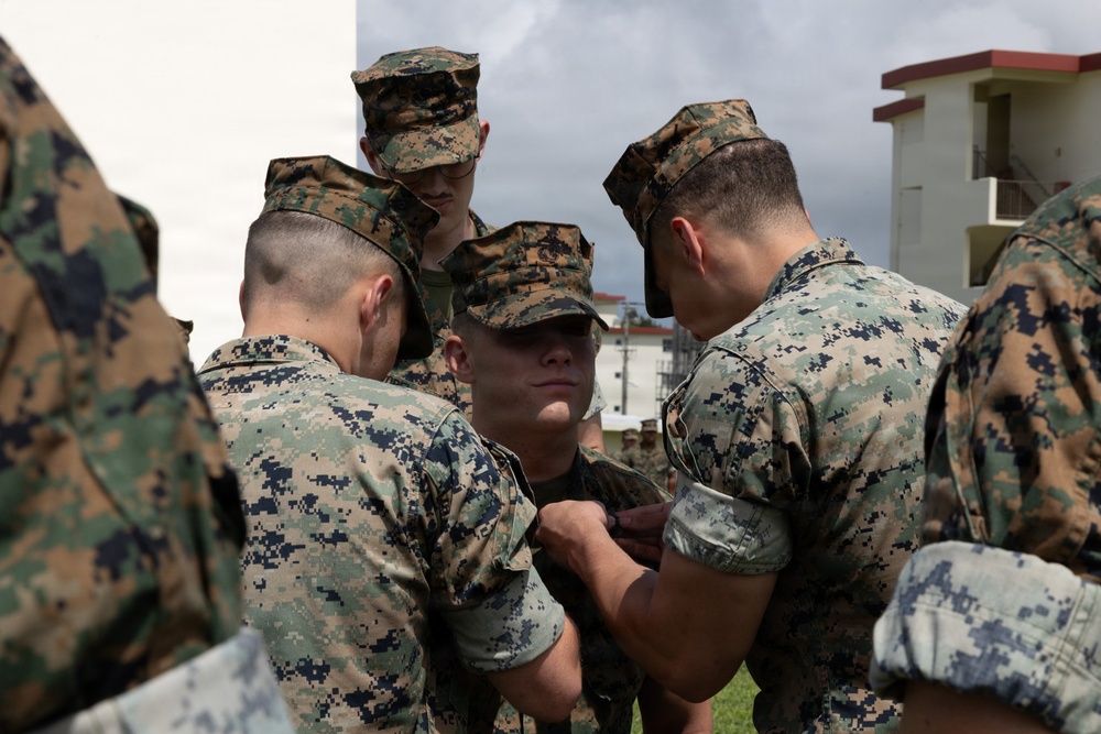 A group of military personnel in digital camouflage uniforms are gathered closely together, appearing to pin something onto the uniform of a central figure. The scene appears to be an informal awards or promotion ceremony taking place outdoors near a building. Multiple service members wearing both MARPAT woodland and Navy Working Uniform (NWU) patterns are present.