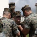 A group of military personnel in digital camouflage uniforms are gathered closely together, appearing to pin something onto the uniform of a central figure. The scene appears to be an informal awards or promotion ceremony taking place outdoors near a building. Multiple service members wearing both MARPAT woodland and Navy Working Uniform (NWU) patterns are present.