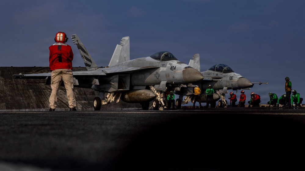 A carrier deck operations scene showing at least two F/A-18 Super Hornet jet aircraft staged on an aircraft carrier flight deck. Deck crew in color-coded jerseys are performing pre-launch or handling procedures around the aircraft. The foreground figure in a red jersey is giving hand signals, consistent with ordnance or aircraft handling roles.