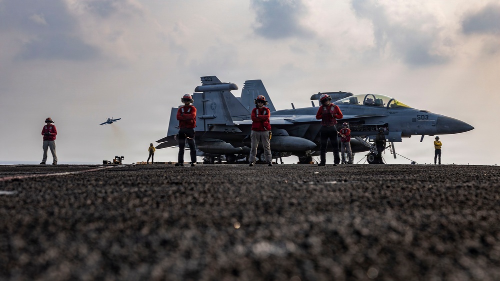 The image shows a U.S. Navy aircraft carrier flight deck with personnel in red jerseys standing near a jet aircraft marked '503', identifiable as an F/A-18E/F Super Hornet or EA-18G Growler. Flight deck crew are positioned around the aircraft in various roles, while another aircraft is visible in the background taking off or flying past. The scene captures routine carrier flight operations under partly cloudy skies.