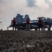 The image shows a U.S. Navy aircraft carrier flight deck with personnel in red jerseys standing near a jet aircraft marked '503', identifiable as an F/A-18E/F Super Hornet or EA-18G Growler. Flight deck crew are positioned around the aircraft in various roles, while another aircraft is visible in the background taking off or flying past. The scene captures routine carrier flight operations under partly cloudy skies.