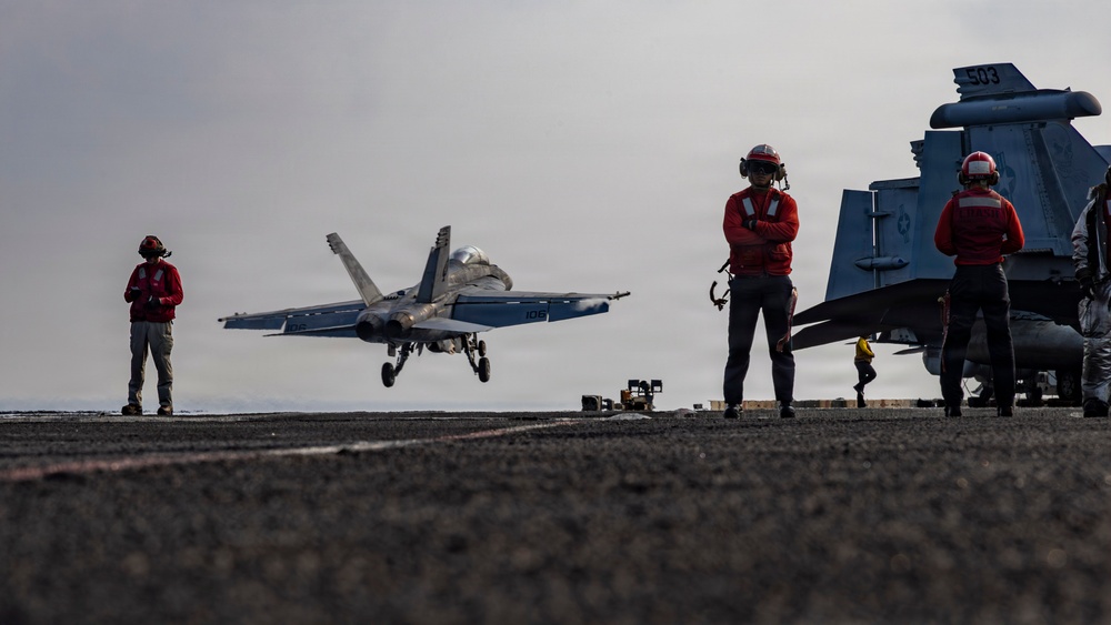 A carrier-based jet aircraft, appearing to be an F/A-18 Hornet or Super Hornet with modex number '106', is captured during a arrested landing or touch-and-go on what appears to be a naval aircraft carrier flight deck. Multiple deck crew members wearing red helmets and jerseys are positioned on the deck observing the operation. A second aircraft with the number '503' is visible parked on the right side of the frame.
