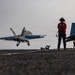 A carrier-based jet aircraft, appearing to be an F/A-18 Hornet or Super Hornet with modex number '106', is captured during a arrested landing or touch-and-go on what appears to be a naval aircraft carrier flight deck. Multiple deck crew members wearing red helmets and jerseys are positioned on the deck observing the operation. A second aircraft with the number '503' is visible parked on the right side of the frame.