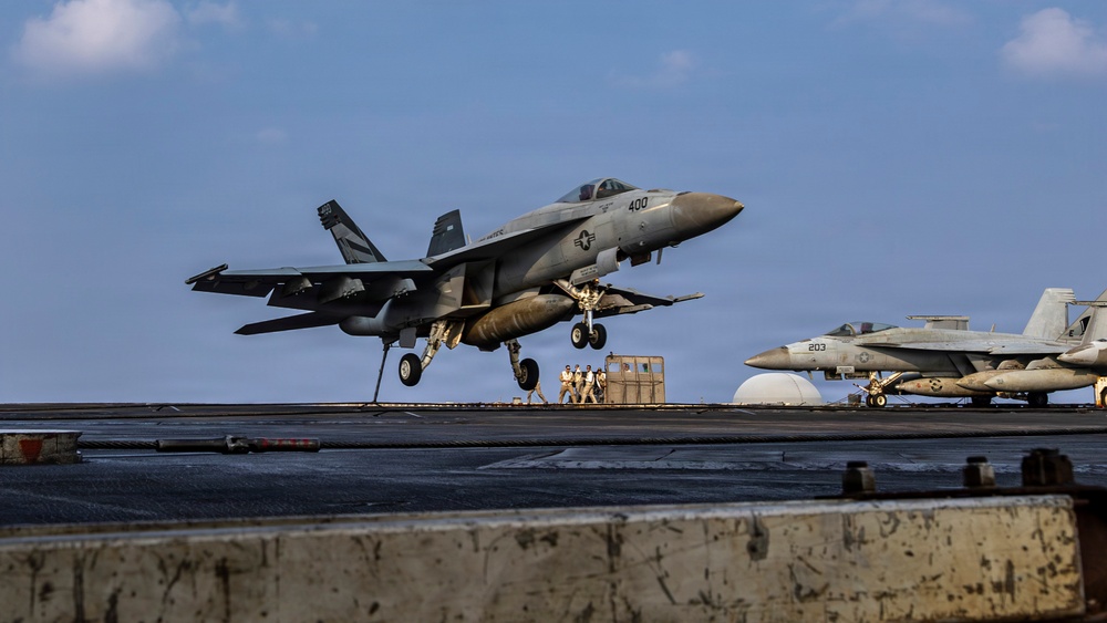A U.S. Navy F/A-18 Super Hornet (bureau number 400) is captured mid-launch or landing on an aircraft carrier flight deck. A second F/A-18 with modex number 203 is visible parked on the deck in the background. Flight deck personnel in colored jerseys are visible near the aircraft.