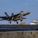 A U.S. Navy F/A-18 Super Hornet (bureau number 400) is captured mid-launch or landing on an aircraft carrier flight deck. A second F/A-18 with modex number 203 is visible parked on the deck in the background. Flight deck personnel in colored jerseys are visible near the aircraft.