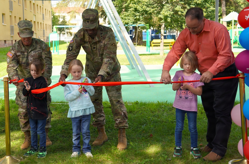 A ribbon-cutting ceremony is taking place at what appears to be a newly opened playground on a military installation. Two U.S. Army soldiers in camouflage uniforms and a civilian man in a red shirt are assisting three young children in cutting a red ribbon. The setting includes playground equipment, colorful balloons, and green grass in the background.