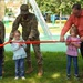A ribbon-cutting ceremony is taking place at what appears to be a newly opened playground on a military installation. Two U.S. Army soldiers in camouflage uniforms and a civilian man in a red shirt are assisting three young children in cutting a red ribbon. The setting includes playground equipment, colorful balloons, and green grass in the background.