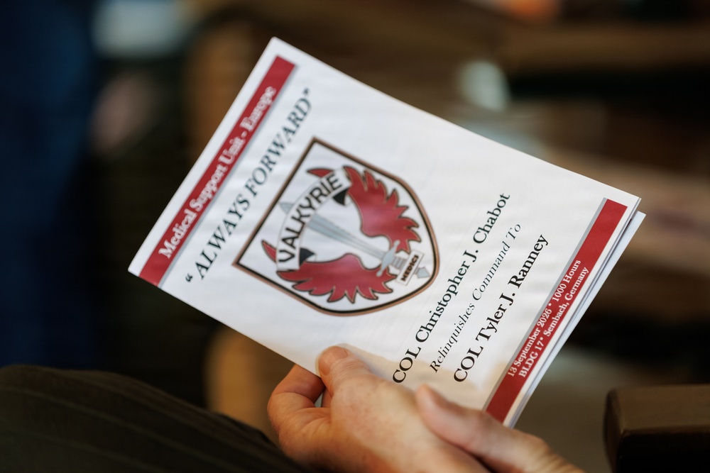 A hand holds a military ceremony program for the Medical Support Unit - Europe, featuring the 'Valkyrie' unit insignia. The program documents a change of command ceremony where COL Christopher J. Chabot relinquishes command to COL Tyler J. Ranney. The event took place on 13 September 2026 at 1000 Hours, BLDC 17, Sembach, Germany.